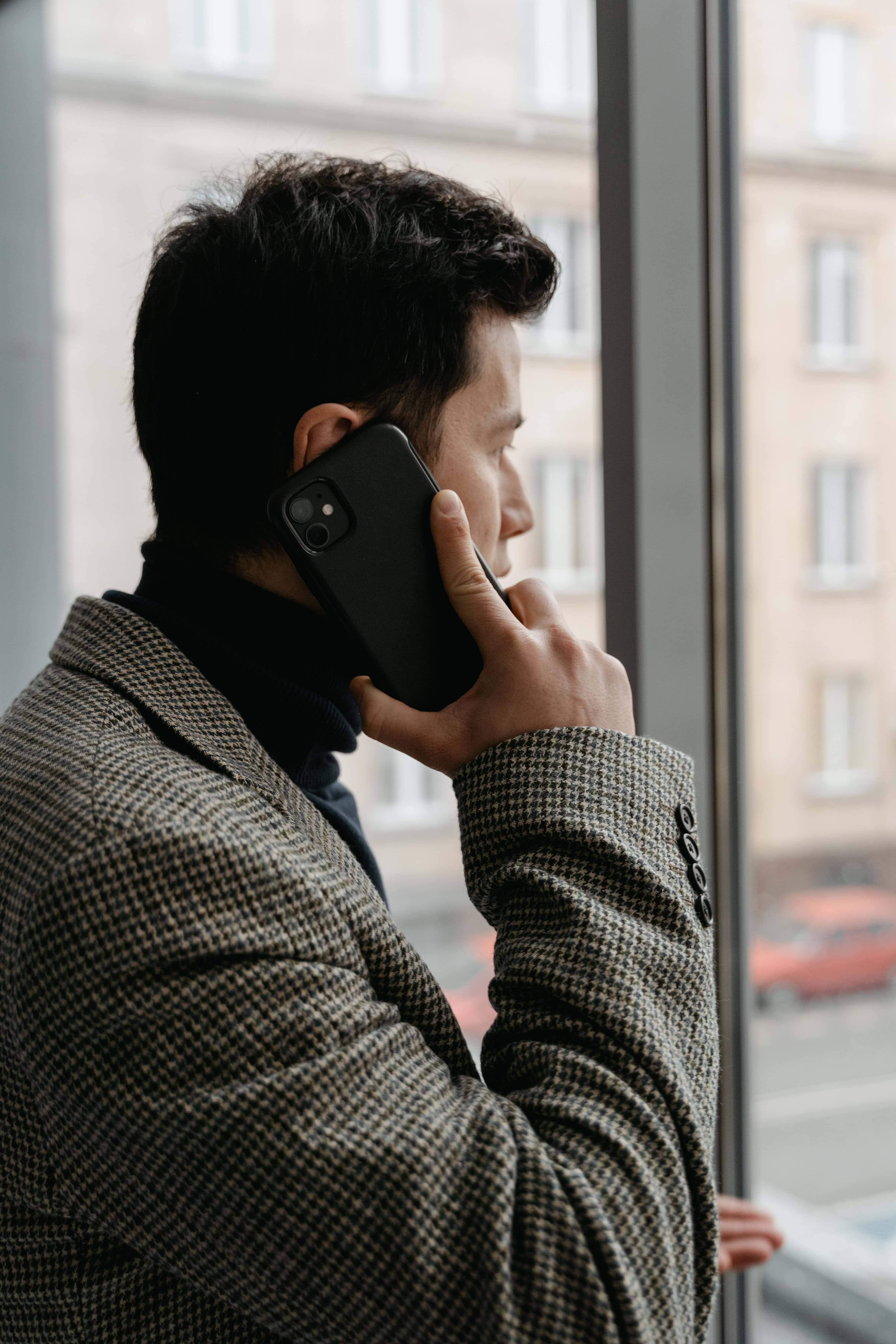 Man in a checkered blazer using a smartphone while gazing out a window