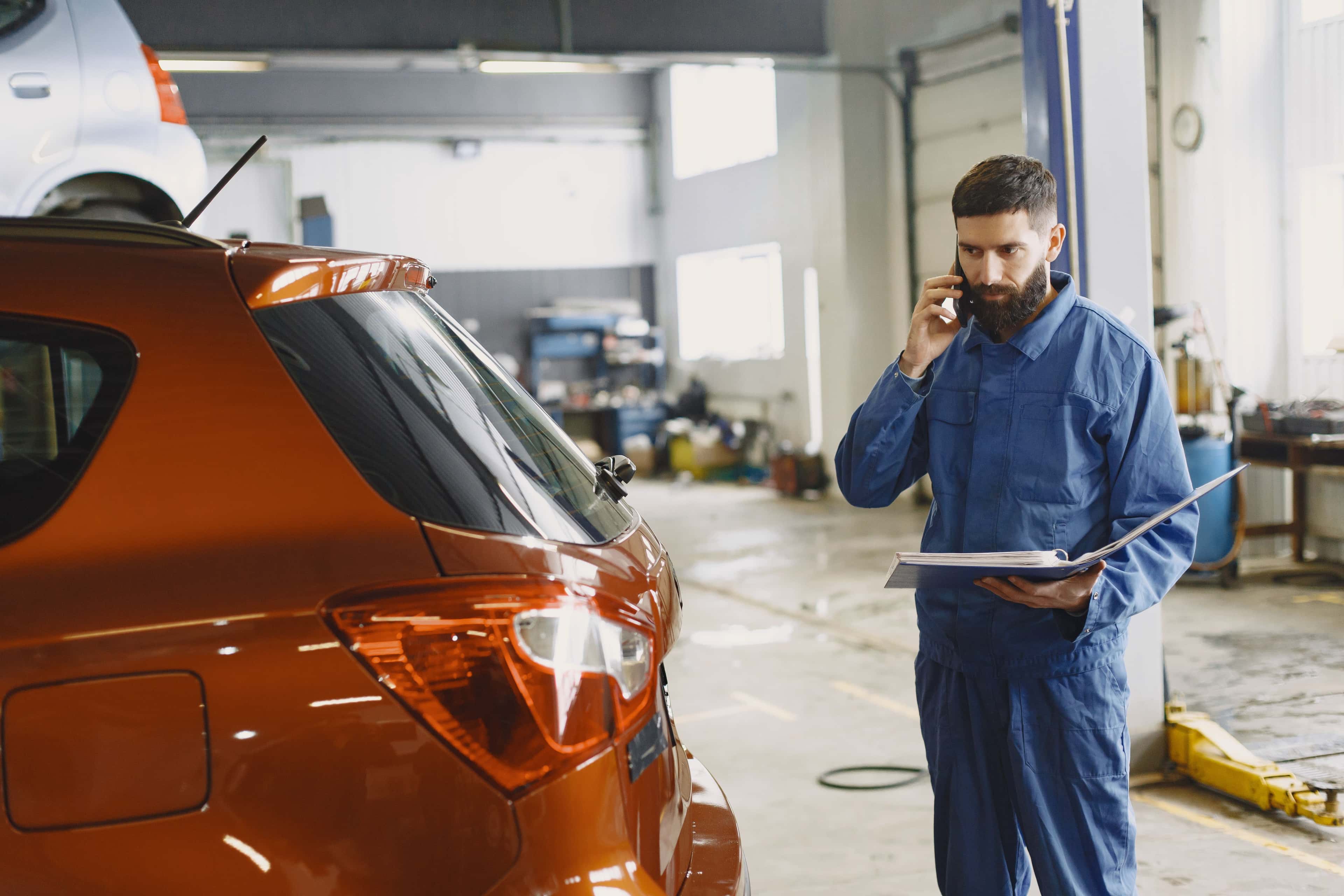 Mechanic in a blue uniform talking on the phone beside a red car in an auto repair shop